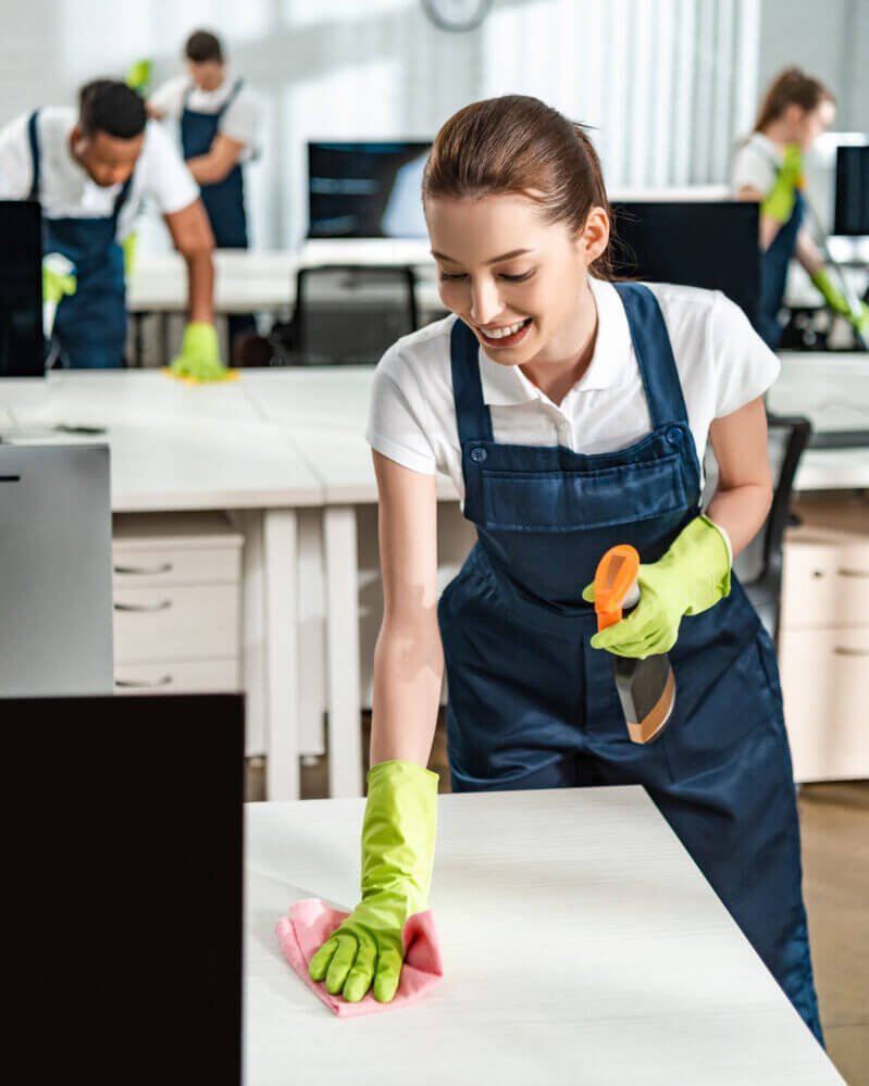 cheerful cleaner in overalls cleaning office desk with rag